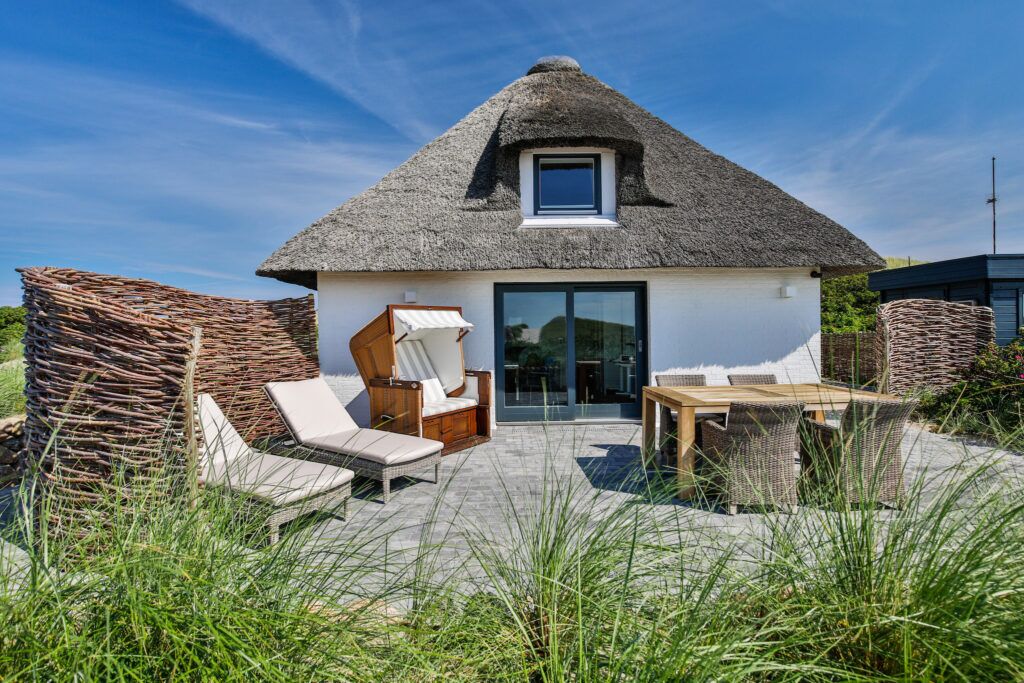 Ein Garten von einem Ferienhaus mit Reetdach, Strandkorb und zwei liegen, Für mehr Privatsphäre erstreckt sich links halb rund ein handgeflochtener Holzzaun.